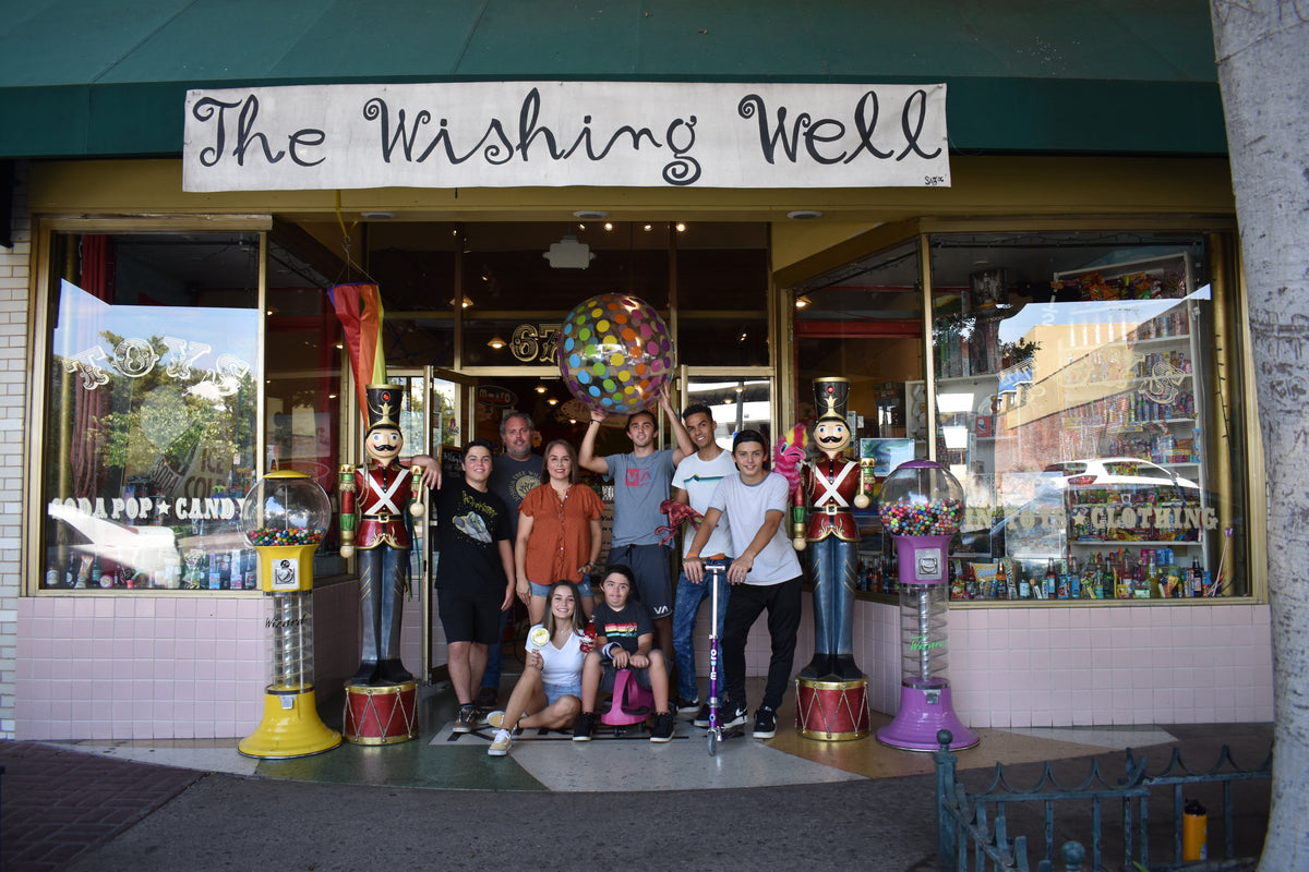 The Wishing Well toy store owner with her children and family posing outside the shop, surrounded by colorful candy machines, toy soldiers, and a large patterned ball.