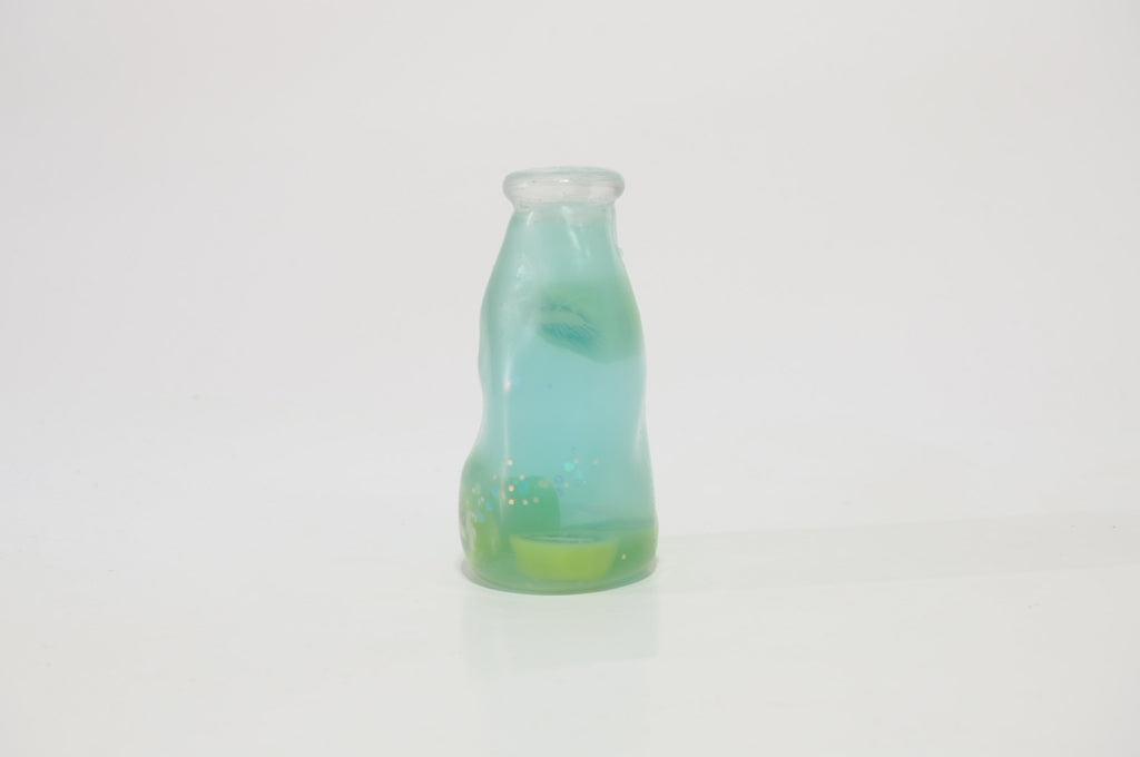 Translucent aqua-blue oil-filled squishy toy shaped like a small fruit drink bottle, with green accents and fruit-shaped pieces visible inside, standing upright against a white background.