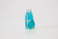 Translucent blue oil-filled squishy toy shaped like a small drink bottle, with fruit-shaped pieces visible inside, standing on a white surface.