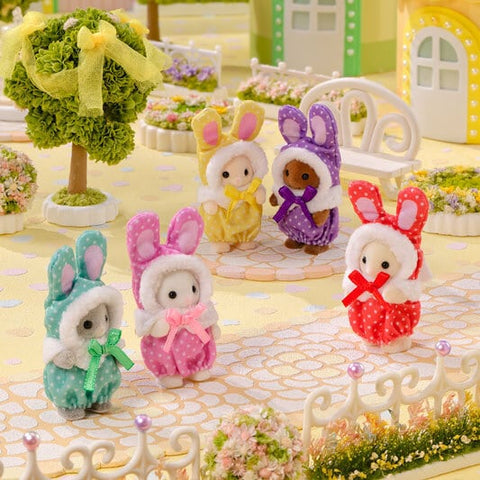 Five Calico Critters baby figures in mint green, pink, yellow, purple, and red bunny costumes arranged in a miniature garden with flowers, decorative fencing, and pastel buildings.
