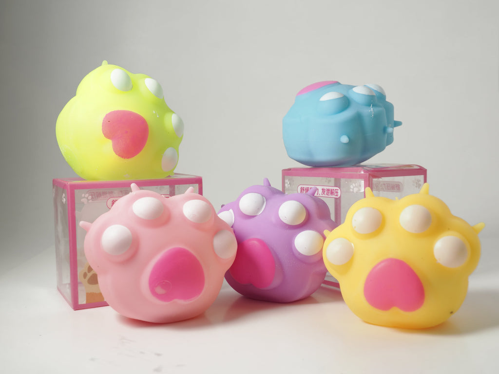 Five cat paw marshmallow squishy toys in green, blue, pink, purple, and yellow, featuring white toe pads and large pink paw pads, displayed with two pink retail boxes.