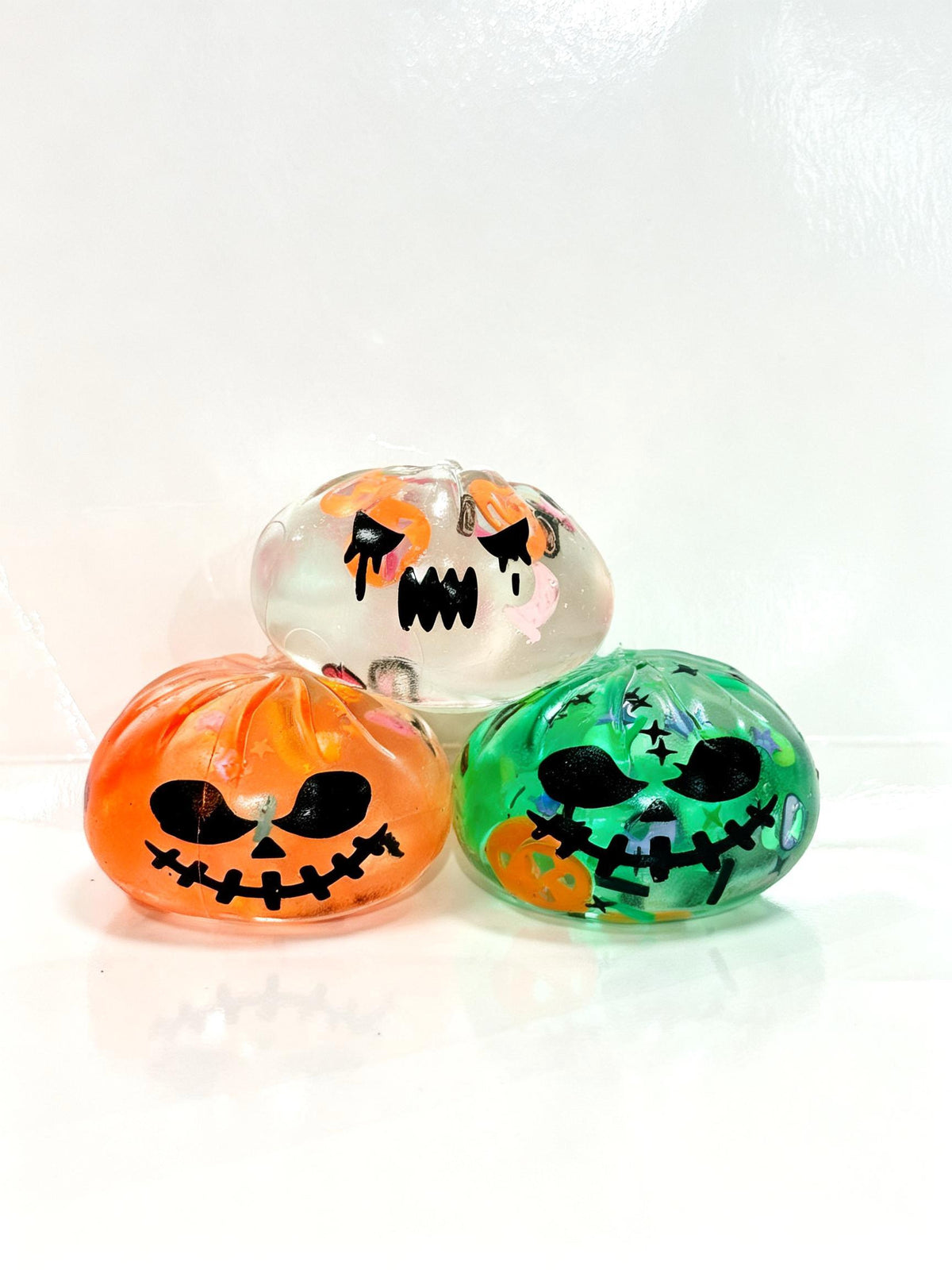 Three decorative translucent pumpkin-shaped ornaments on a white reflective surface against a white background. One is orange with a black stitched jack-o'-lantern face, one is green with a black stitched jack-o'-lantern face and small orange details, and one is clear with orange and black facial details.