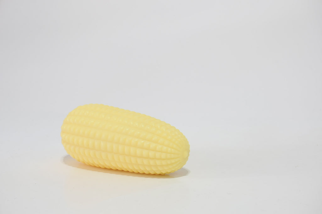 Yellow corn-shaped crunchy squishy toy resting diagonally on a white surface, photographed from an angled side view.