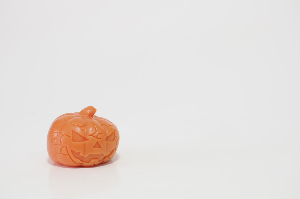 Orange jack-o'-lantern crunchy squishy photographed at an angle, showing its mischievous face, triangular nose, jagged smile, rounded pumpkin shape, and short stem.