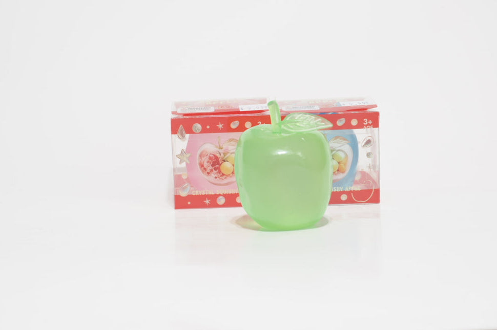 Translucent light green crystal apple oil squishy with a matching stem and leaf, standing in front of red-trimmed retail packaging on a white surface.