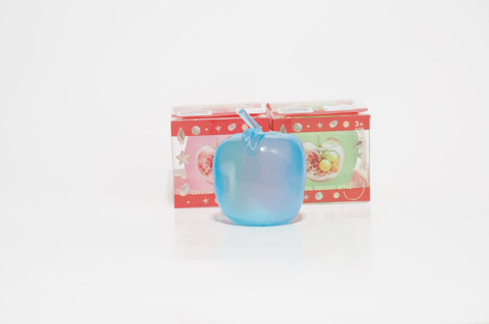 Translucent turquoise-blue crystal apple oil squishy with a matching stem and leaf, standing in front of two colorful red-bordered retail boxes on a white surface.