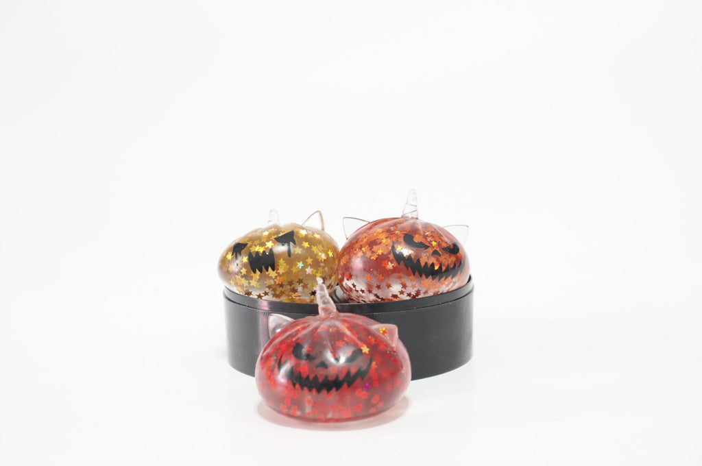 Three Halloween Spooky Triplets Dumplings squishy toy designs with glittery star patterns and black jack-o'-lantern faces. Yellow and orange designs sit on a black circular stand, with a pink-red design displayed in front.