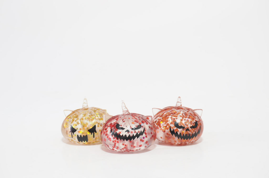 Front view of three Halloween Spooky Triplets Dumplings squishy toy designs: a yellow one with a frowning face, a pink-and-white speckled one with a jagged grin, and an orange-and-white speckled one with angry eyes.