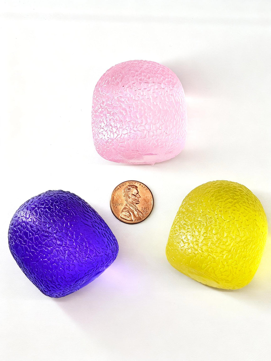 Pink, purple and yellow gumdrop-shaped squishies arranged around a penny for size comparison.