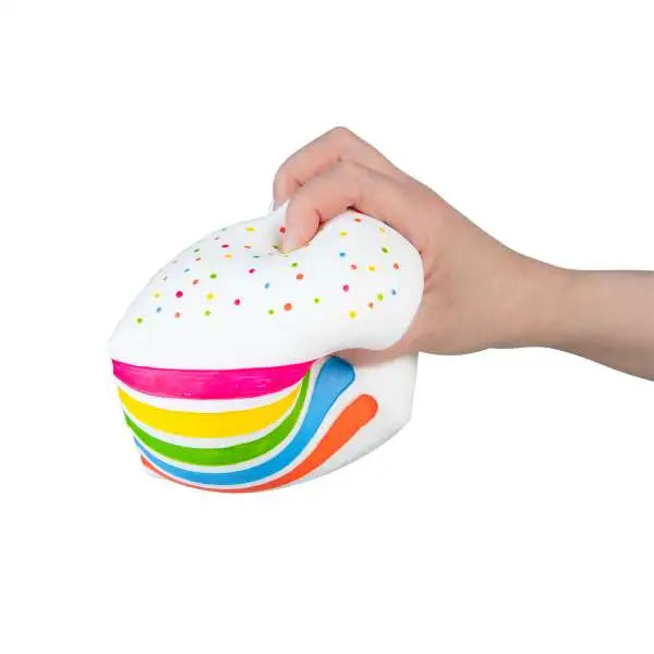 A hand squeezes the white frosted top of a rainbow cake-slice squishy, bending its colorful layers.