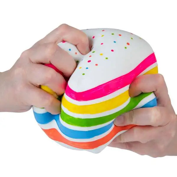 Two hands compress a rainbow cake-slice squishy with white frosting, colored sprinkles and bright horizontal layers.