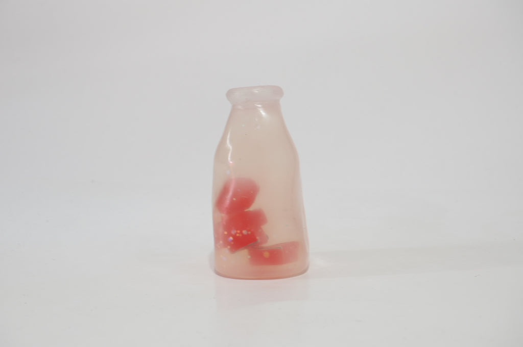Translucent pale pink fruit drink bottle-shaped oil squishy with small red fruit-shaped pieces inside, standing upright against a white background.